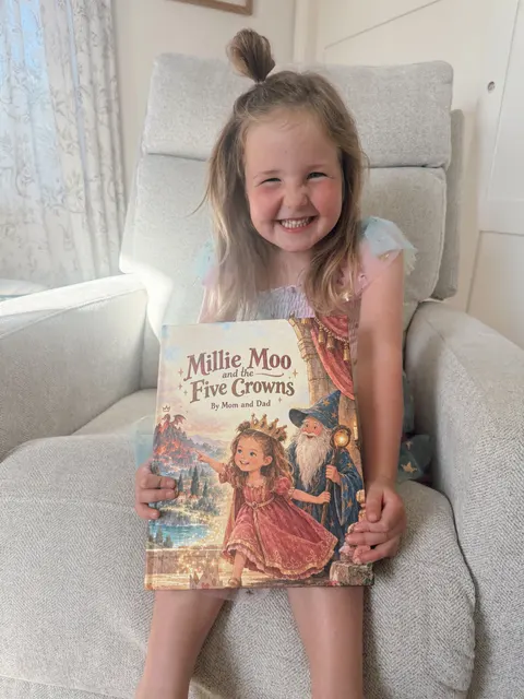A smiling child holding Millie Moo and the Five Crowns in an armchair