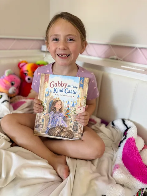 A smiling child holding Gabby and the Kind Castle on a bed