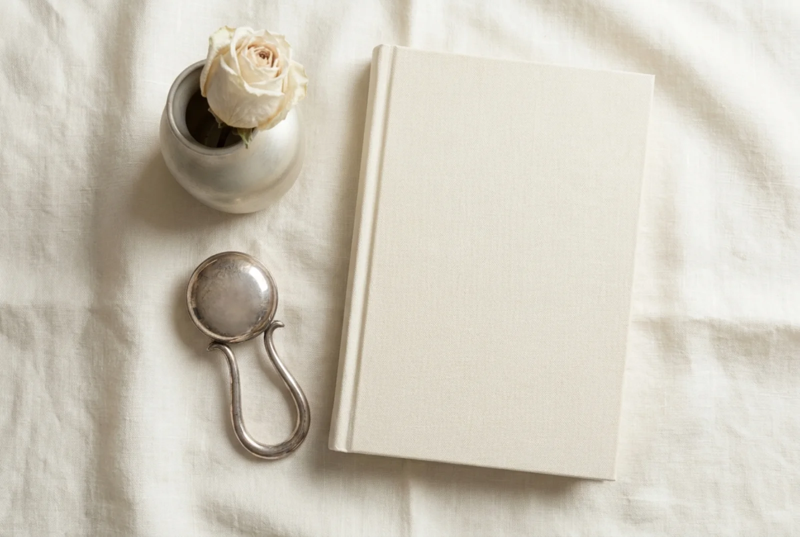 A small silver christening cup on a white linen runner beside a cream linen hardcover book and a folded white knitted blanket in soft window light against pale wood.