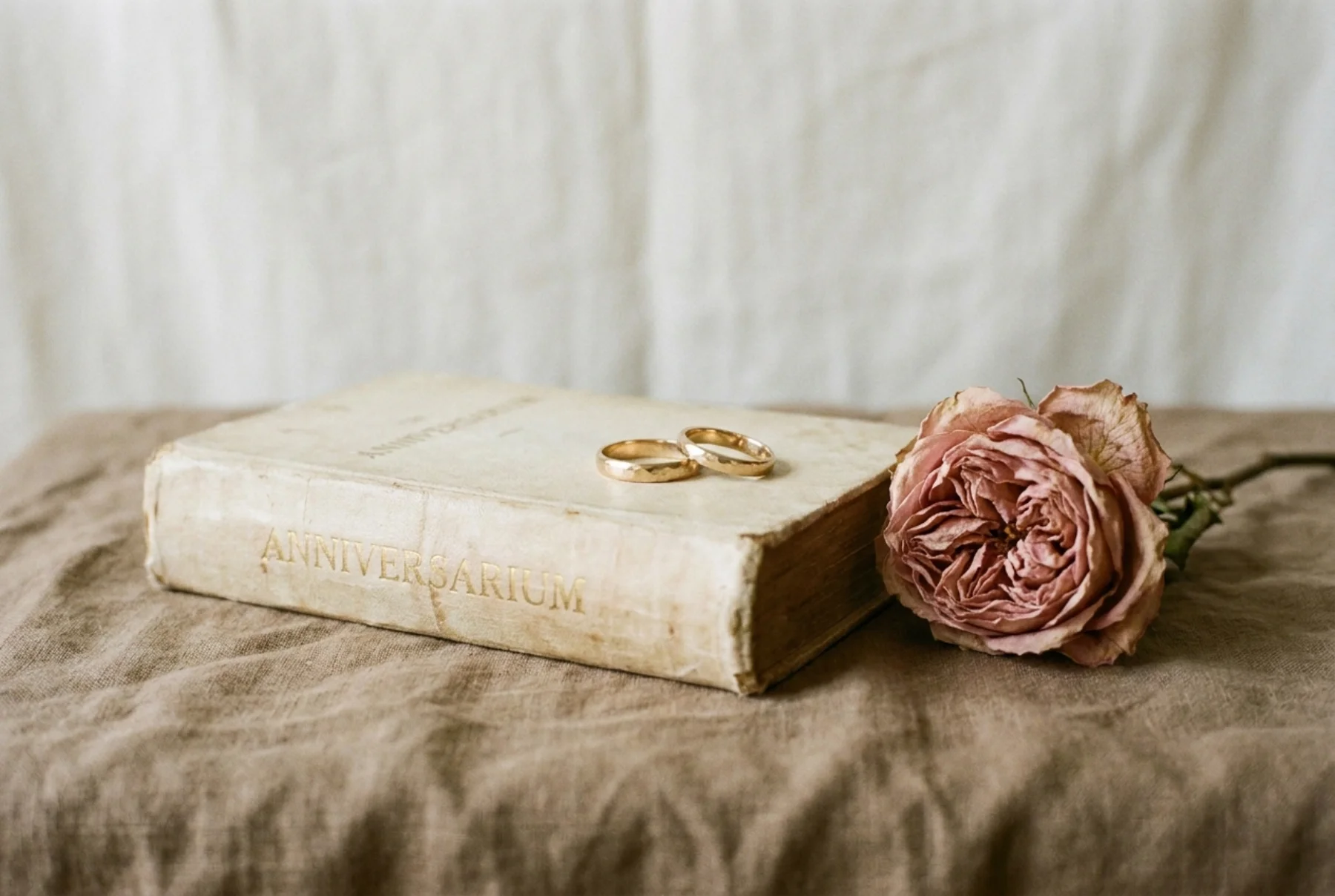 A foil-stamped hardcover golden anniversary keepsake book and a small pair of gold rings resting on warm linen in soft window light.