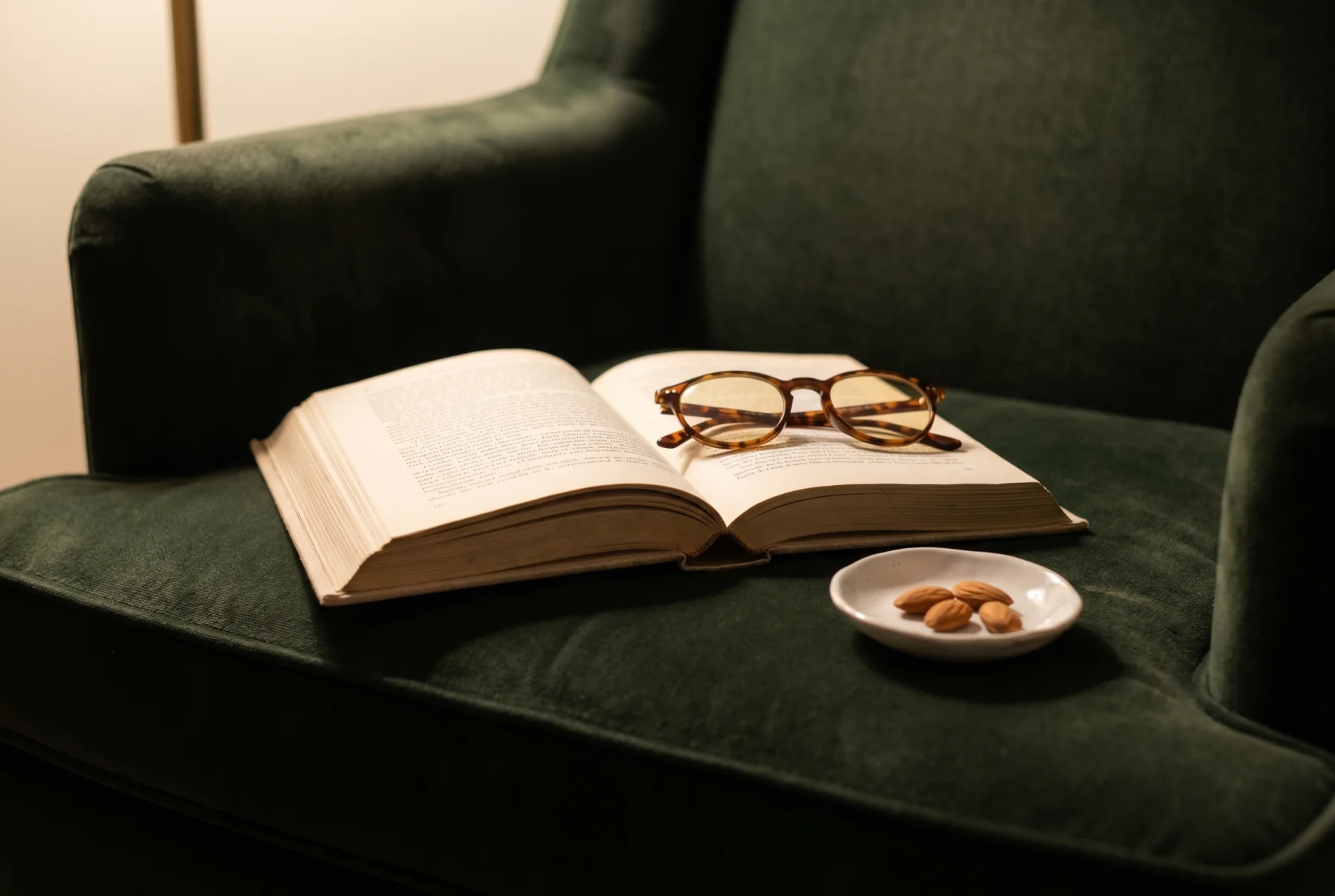 A cream hardcover book lying open on a forest-green velvet armchair, tortoiseshell reading glasses on the right page, a porcelain dish of almonds alongside, warm lamp light.