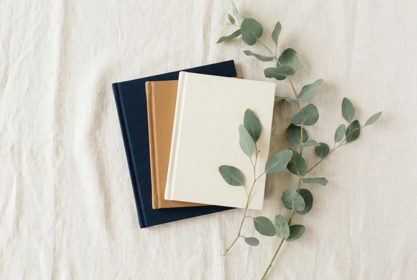 A cream linen hardcover book lying flat on a pale oak nursery shelf beside a small wooden figure and a folded muslin blanket, north-window light falling across the cover.