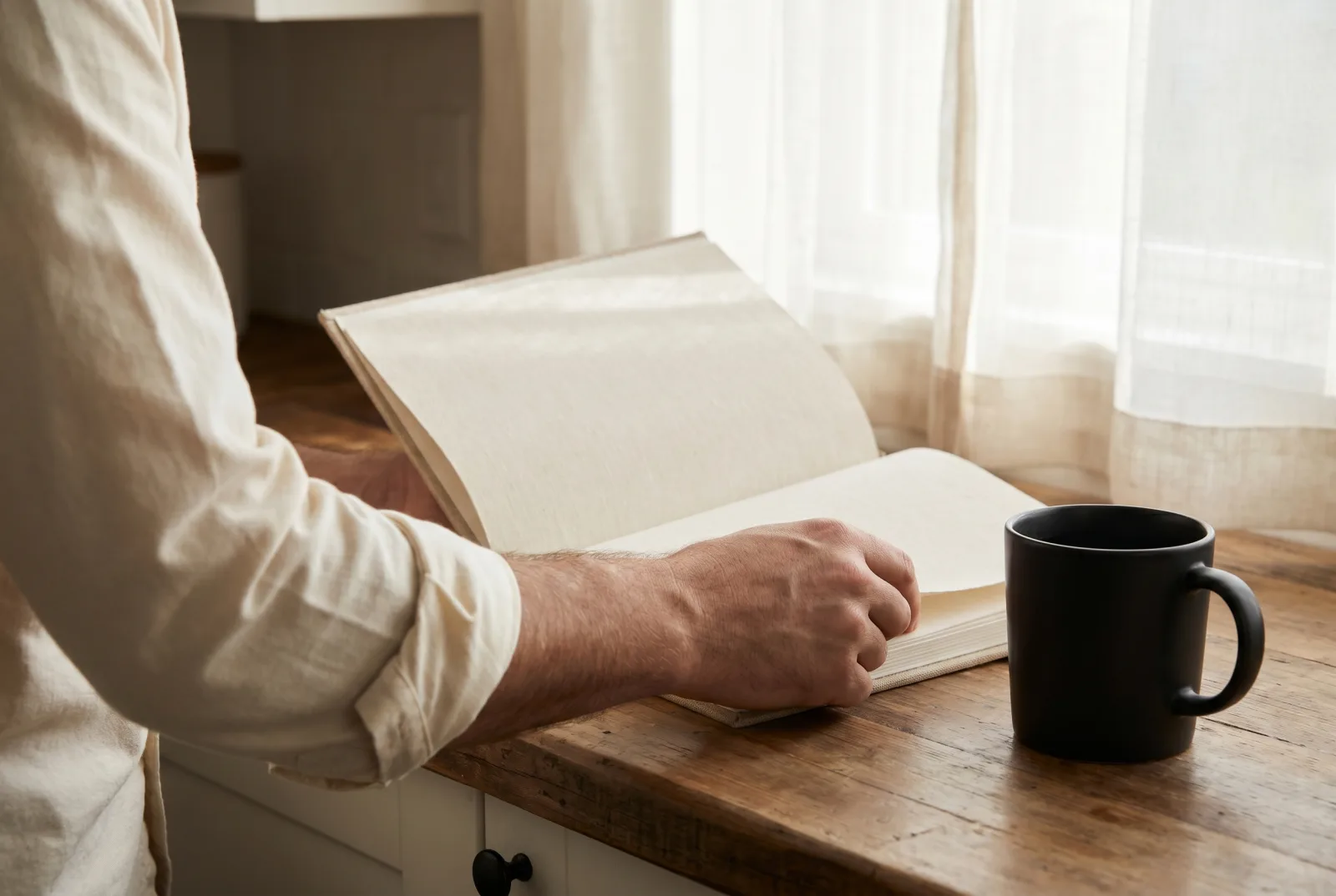 A linen hardcover book open to a blank page held against a man's forearm in a rolled cream shirt cuff on a kitchen counter beside a matte black coffee mug.